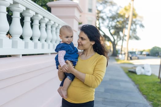 Portrait of happy mother holding her cute baby outside white and pastel-pink building. Beautiful brunette woman and her adorable young daughter in peaceful neighborhood. Lovely family outdoors