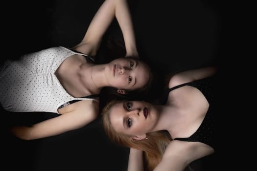 Pair of young women is lying on the black floor looking at the camera. Horizontally.