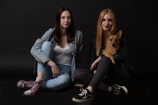 Pair of young women is sitting on the black floor holding brown dog.  Horizontally.