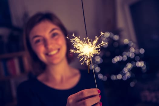 Smiling girl is holding a sparkler in her hand, indoor, lights in background