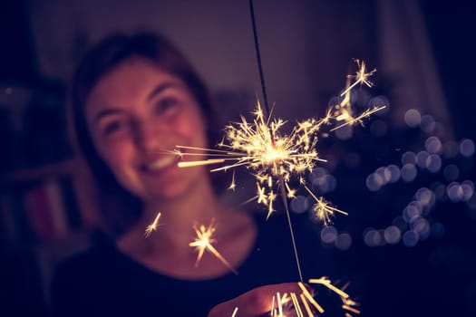 Smiling girl is holding a sparkler in her hand, indoor, lights in background