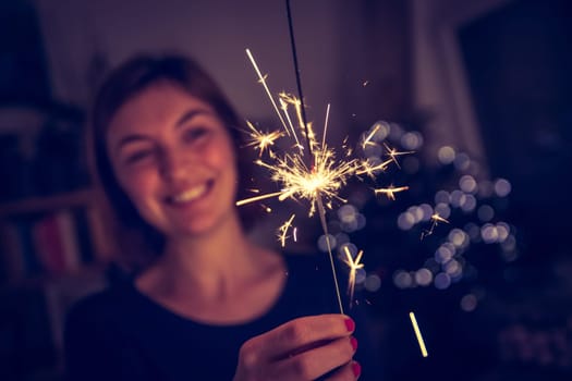 Smiling girl is holding a sparkler in her hand, indoor, lights in background