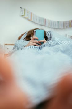 Girl lying in bed is using her smartphone, smartphone in the foreground, blurry background
