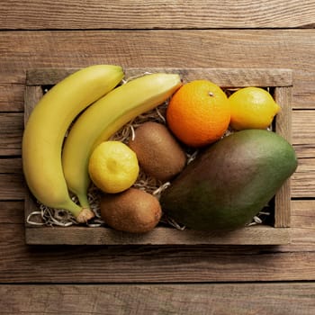 Fresh tropical fruits in a wooden delivery box on a wooden background. Orange, banana, mango, kiwi and lemon top view flat lay with copy space for some advertisement text