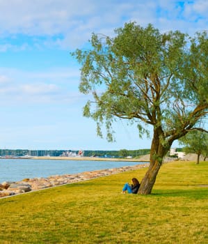 Woman read a book under the tree in a park near the sea. Tallinn, Estonia