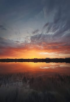 Fascinating sunset reflecting on the lake surface. Idyllic landscape, vertical background. Silent and tranquil evening scene with colorful sky, reed vegetation in the pond and a city at the horizon