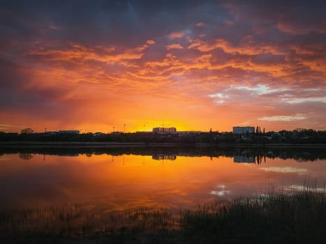 Colorful and dramatic sunset sky reflecting on pond water. Silent evening scene at Delia lake. Vibrant sundown clouds at the horizon over Ungheni city, Moldova. Reed and rush vegetation in the water