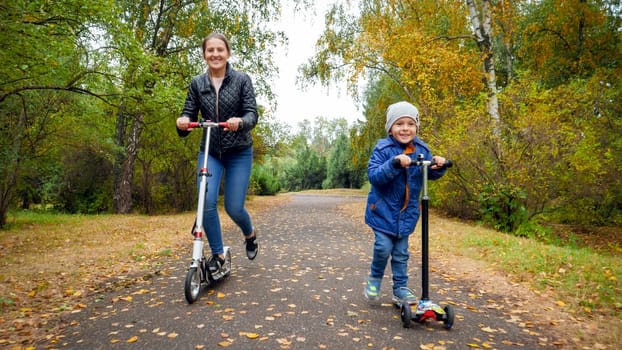 Happy smiling boy with mother racing on kick scooters at autumn park.