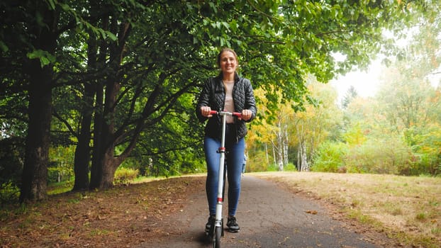 Beautiful smiling young woman riding on kick scooter at autumn park.