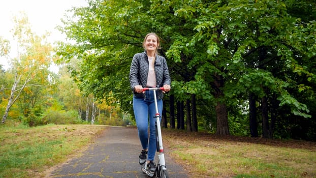 Happy young woman riding on scooter under big trees at autumn park.