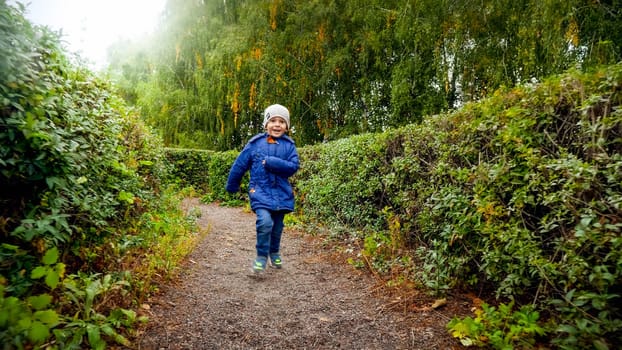 Happy laughing little boy in jacket running in bush labyrinth at autumn park.