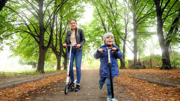 Happy smiling little boy with young mother riding on scooters on long tree alley at autumn park.