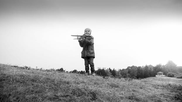 Black and white image of smiling little boy standing on hill top and throwing toy aitplane.