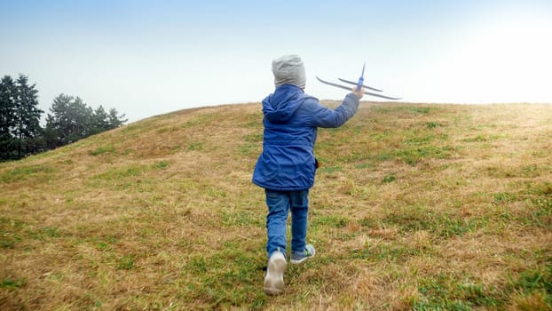 Rear view image of little boy running with toy airplane on the hill top.