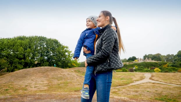 Portrait of happy smiling young mohter holding and embracing her little son on field at park.