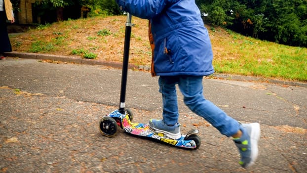 Closeup photo of little boy standing and riding on kick scooter at autumn park.