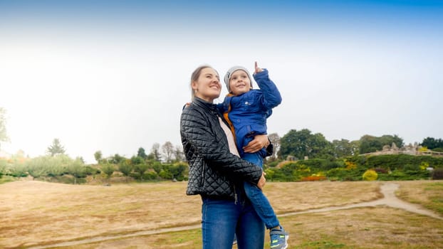 Portrait of little boy hugging mother and pointing with finger in the air at park.