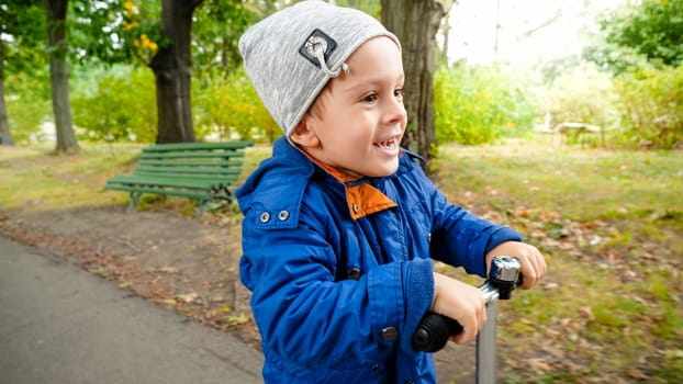 Portrait of smiling toddler boy riding fast on scooter at autumn park.