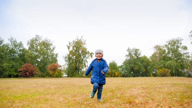 Happy smiling toddler boy in jacket running on field with dry grass on cloudy autumn day.