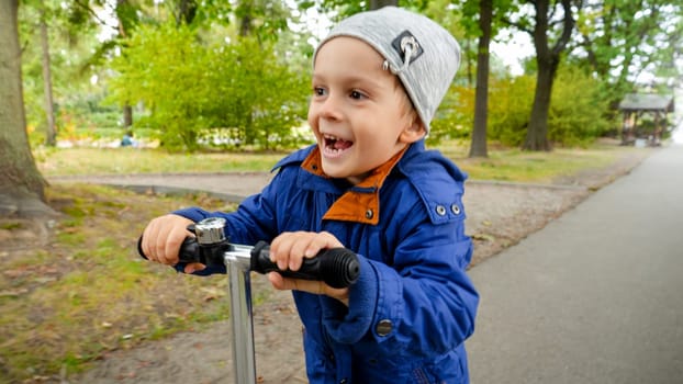 Portrait of laughing and smiling little boy holding steering handle on kick scooter while riding in park.