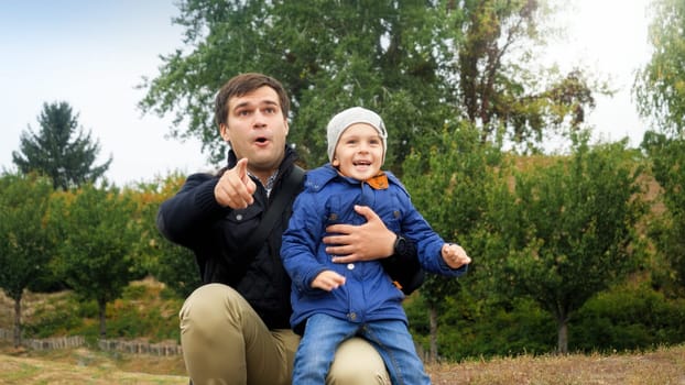 Portrait of amazed smiling father with little boy looking in the air and pointing with fingers at park.
