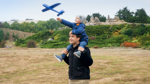 Smiling little boy sitting on fathers shoulders and throwing toy airplane at park.