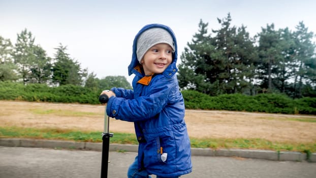 Portrait of smiling little boy in jacket riding on scooter in park.