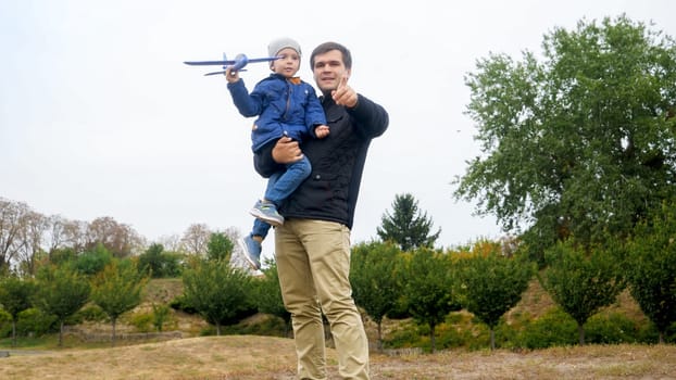 Portrait of young father holding his little son and teaching throwing toy airplane at park.