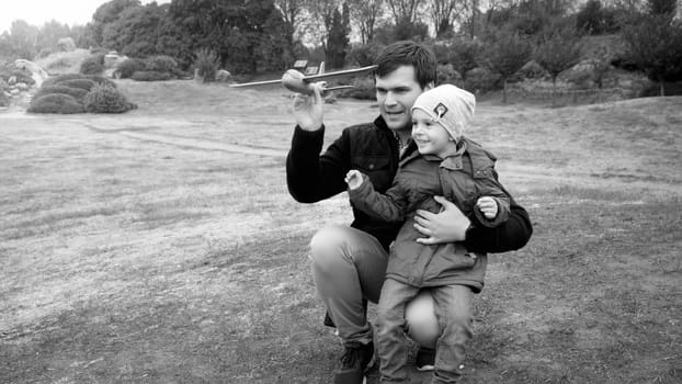Black and white portrait of young father with little son throwing toy airplane on the field at park.