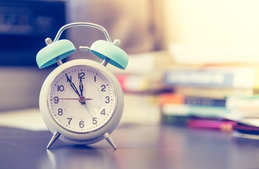 Alarm clock is standing on a grey desk, books and office stuff in the blurry background