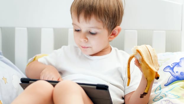 Portrait o flittle toddler boy having snack and playing games on digital tablet computer in bed.