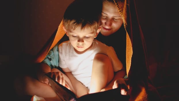 TOned portrait of smiling mohter with little boy sitting under blanket at bedroom and reading book with torch.