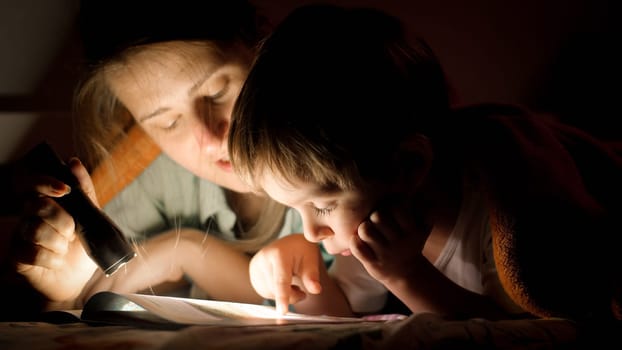 Closeup portrait of young mother holding flashlight while reading a book to her little son at night.