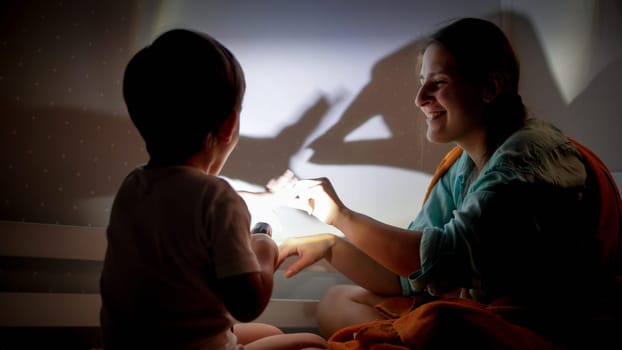 Portrait of smiling young mother and little boy playing with flashlight and shadows on wall at night.