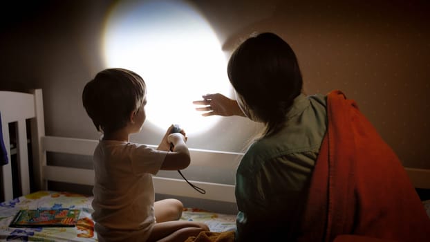Little boy and young mother showing shadow theater on wall in bedroom at night.