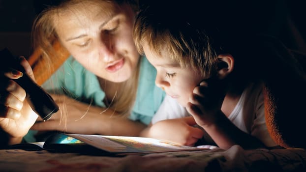 Toned portrait of young mother holding flashlight and reading bedtime story book to her little son lying under blanket at night.