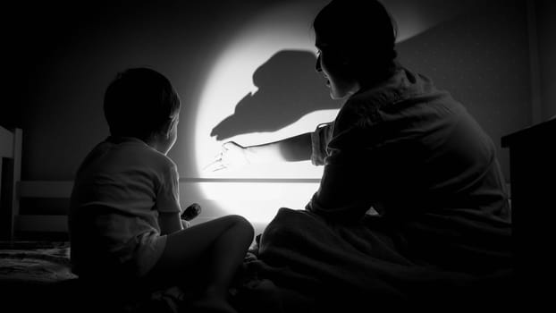 Black and white photo of mother and her child playing with shadows and flashlight on wall at bedroom.