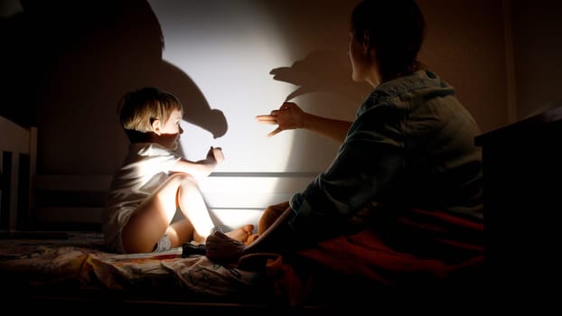 Image of little boy with young mother showing play in theater of shadows on wall at dark room.