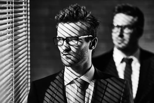 Elegant young businessman in eyeglasses looking through jalousie on background of colleague