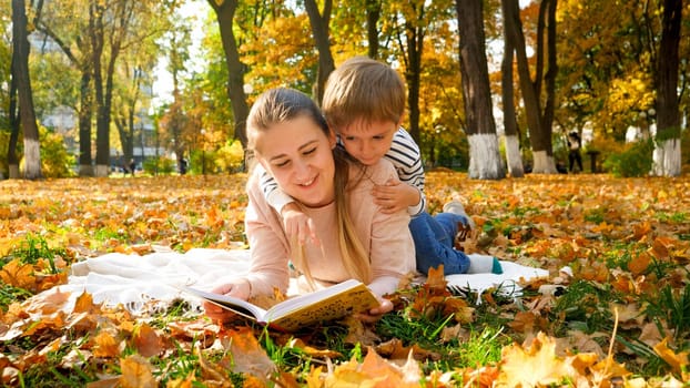 Adorable toddler boy sitting on mothers back and reading a book at autumn park.