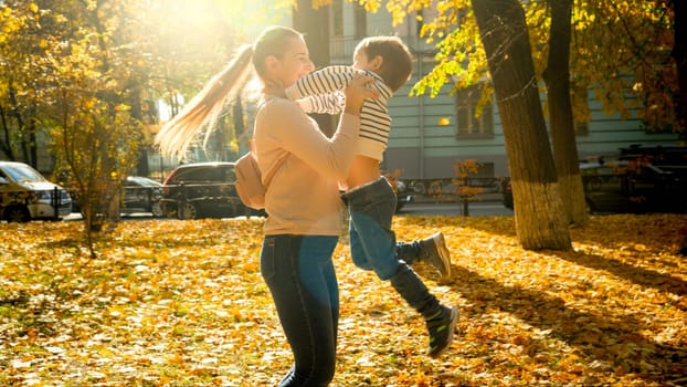 Beautiful happy mother hugging and spinning her laughing little son in autumn park.