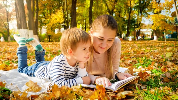 Portrait of little boy lying with his mother at autumn park and reading a book.