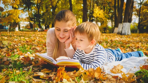 Toned portrait of young mother reading story book to her son lying on autumn leaves at park.
