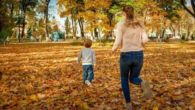 Happy young mother with her little son running on fallen yellow leaves in autumn park.