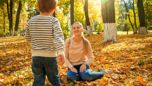 Little boy walking to his mother sitting on autumn leaves at park.
