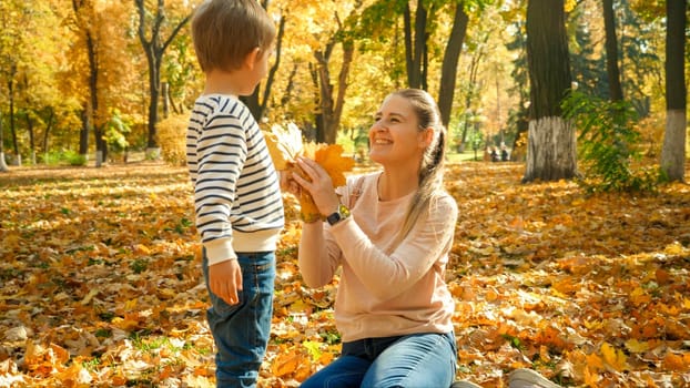 Hapyy smiling boy giving beautiful yellow and red leaves to his mother in autumn park.