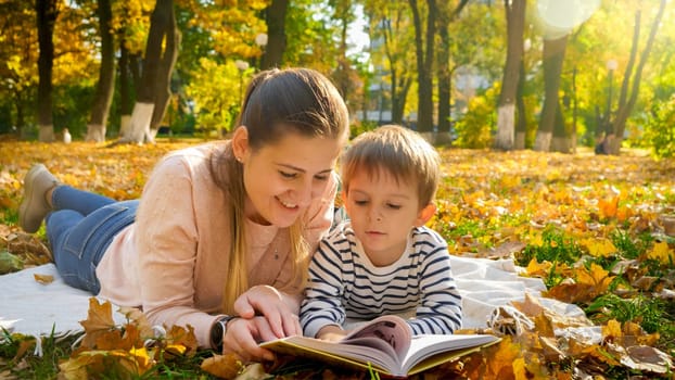 Portrait of beautiful young woman with her little son lying in autumn park and reading a book.