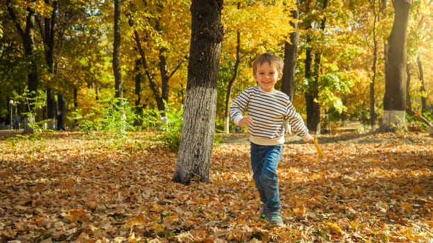Happy laughing toddler boy running over fallen autumn leaves at park.