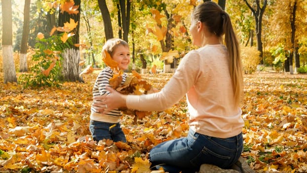 Happy laughing boy with young mother sitting on ground at park and throwing up autumn leaves.