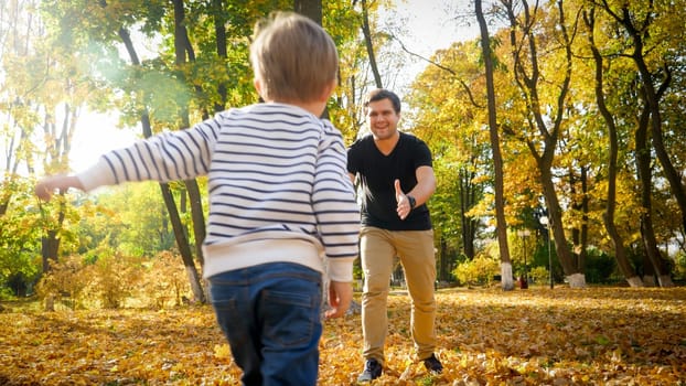 Little toddler boy running to his smiling father at autumn park.